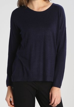 Femme portant un pull bleu marine à manches longues et col rond et un pantalon noir, debout devant un fond clair uni.
