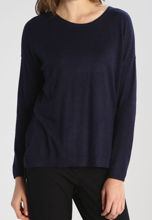Femme portant un pull bleu marine à manches longues et col rond et un pantalon noir, debout devant un fond clair uni.