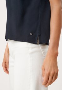 Nahaufnahme einer Person in einem marineblauen Oberteil mit seitlichem Schlitz und weißen Hosen, deren rechte Hand entspannt an der Seite hängt.