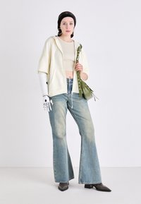 Hvit hettegensere over en beige crop top, blåstripete vide jeans, svarte hælboots og en grønn veske med frynser. En robotarm synlig.