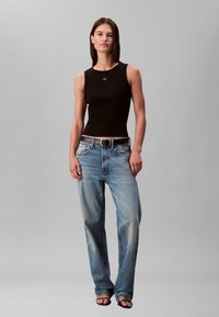 Vrouw draagt een nauwsluitende zwarte mouwloze top, lichtblauwe verwassen jeans met een riem, en zwarte sandalen met open neus, staand tegen een effen grijze achtergrond.
