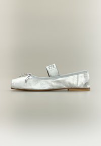 Ασημί μεταλλικό μπαλαρίνα με τετράγωνη μύτη, μικρό φιόγκο μπροστά και ελαστικό λουράκι με εκτυπωμένη ετικέτα μεγέθους.