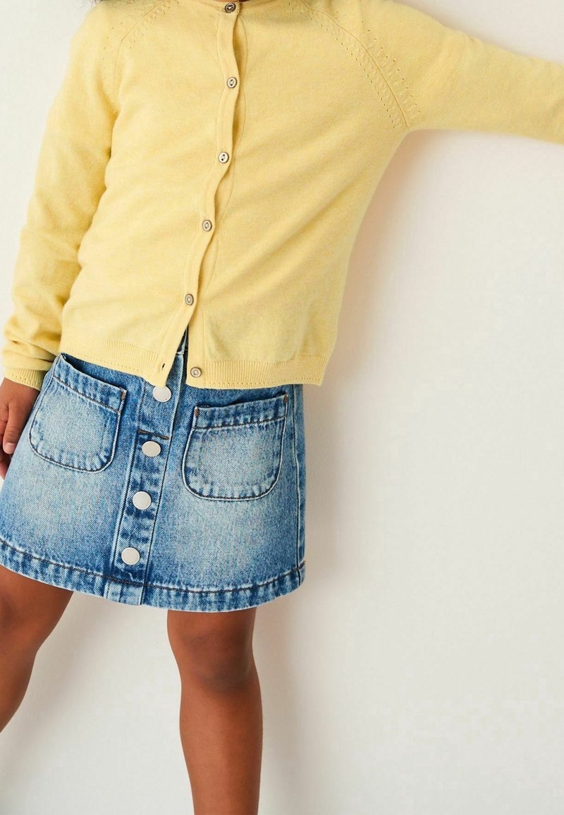 Enfant portant un cardigan jaune boutonné et une jupe en denim bleue avec boutons et poches à l'avant, debout devant un fond clair uni.