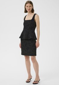 Robe peplum noire avec décolleté carré, texture florale, coupe ajustée, jupe haute au genou. Associée à des sandales à talons noires.