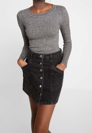 Femme portant un haut gris côtelé ajusté à manches longues et une jupe noire taille haute boutonnée à l'avant, les mains dans les poches.