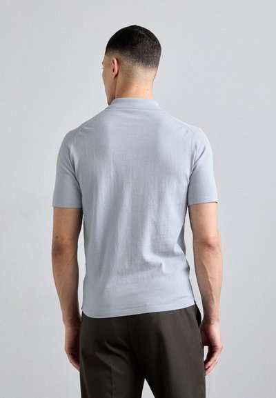 Polo shirt léger bleu à manches courtes avec une texture lisse, un col classique et des fentes latérales, associé à un pantalon sombre.