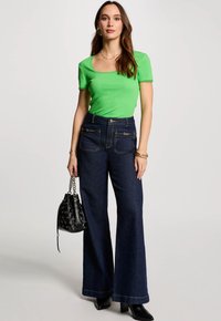 Groene geribbelde top met korte mouwen, hoogwaist donker denim wijde pijpen spijkerbroek met voorkant ritszakken, en zwarte handtas met zilveren accenten.