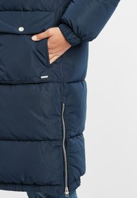 Navy blue padded coat with a zipped side pocket, elastic cuffs, and a snap-buttoned front pocket, featuring a smooth, shiny texture.