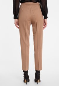 Pantalons beiges ajustés avec une texture lisse, dotés d'une taille haute, de plis centraux et d'une coupe slim. Portés avec des bottines à talons hauts noires.