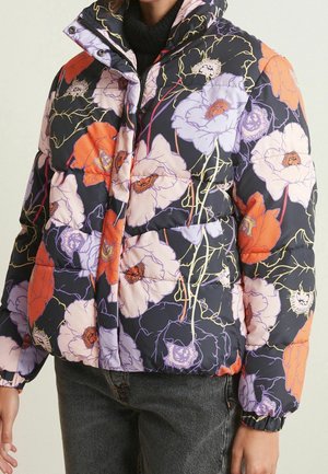 Femme portant une doudoune noire avec un grand imprimé floral orange, violet et blanc, avec des poignets élastiques, associée à un jean foncé.