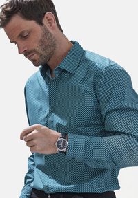 Türkises Button-Down-Hemd mit geometrischem weißen Muster, langen Ärmeln und spitzem Kragen. Eine silberne Uhr mit schwarzem Band ist sichtbar.