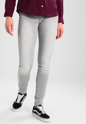 Personne portant un jean skinny gris clair légèrement usé, des baskets noires et blanches, et un t-shirt à manches longues rouge foncé marchant vers l'avant.