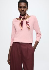 Világos rózsaszín hosszú ujjú felső lekerekített nyakkivágással, melyet mély bordó, magasderekú nadrággal párosítottak. Egy bordó és barna selyemsál egészíti ki az öltözéket.