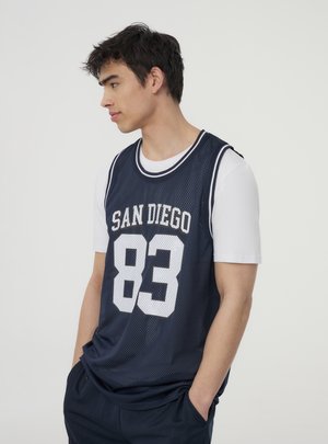 Maglia da basket navy in tessuto mesh con la scritta "SAN DIEGO" e "83" in lettere bianche. Indossata sopra una maglietta bianca, il modello ha le mani nelle tasche.