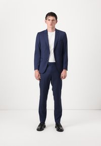 Marineblaues Anzug mit schlanker Passform, einreihigem Sakko und glatten Vorderhosen, kombiniert mit einem weißen T-Shirt mit Rundhalsausschnitt und schwarzen Schuhen.