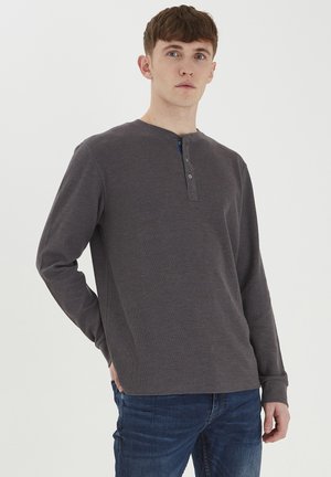 Cămașă henley gri cu mânecă lungă, realizată dintr-un material texturat. Are un decolteu rotund și o închidere cu două nasturi. Este asociată cu blugi din denim.