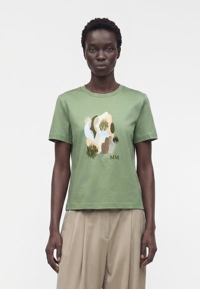 Woman wearing olive green t-shirt with abstract beige, white, and brown shapes and "MM" letters, paired with loose beige pants.