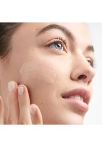 Lichtbeige foundation aangebracht op een lichte huid op de wang; model raakt zachtjes het gezicht aan met natuurlijke nagels en een subtiele lipkleur.