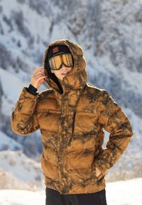 Braun und schwarz gemusterte Daunenjacke mit Kapuze, mit einem Reißverschlusstaschen auf der Brust und elastischen Bündchen, getragen mit einer schwarzen Mütze und Skibrille in einer verschneiten Umgebung.