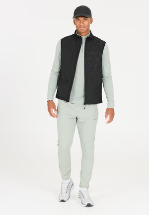 Gilet matelassé noir sur une chemise à manches longues vert clair, associé à un pantalon vert clair et des baskets blanches, avec une casquette noire.