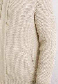 Beige gestrickte Strickjacke mit einem strukturierten Muster, ausgestattet mit einer Fronttasche, Druckknopfverschluss und einer Kapuze mit Kordelzug. Etikett am Ärmel.