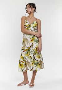 Vestido floral sin mangas con patrones de hojas verdes y amarillas, un corsé ajustado y una falda con volantes. Llevado con sandalias de espuma blanca.