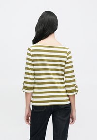 Svītrains krekls zaļā un baltā krāsā ar plašu kakla izgriezumu un trīs ceturtdaļu piedurknēm ar pogu dekorācijām. Kombinēts ar tumšām džinsu biksēm.