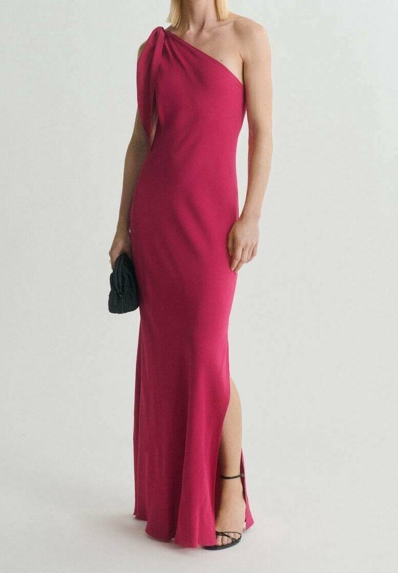 Femme portant une robe fuchsia longue jusqu’au sol, asymétrique avec une fente latérale, tenant une pochette noire et portant des talons noirs à brides.