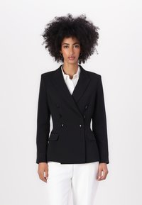 Blazer preto de dupla abotoadura com lapelas em pico, dois bolsos frontais e botões pretos brilhantes, combinado com uma camisa branca e calças claras.