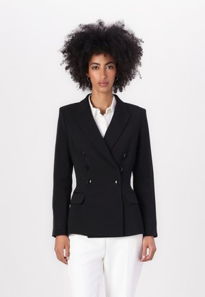 Blazer nero a doppio petto con revers a punta, due tasche frontali e bottoni neri lucidi, abbinato a una camicia bianca e pantaloni chiari.