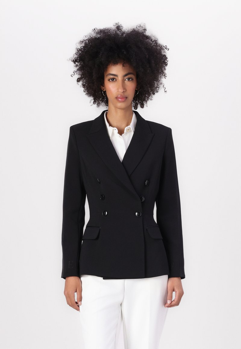 Blazer preto de dupla abotoadura com lapelas em pico, dois bolsos frontais e botões pretos brilhantes, combinado com uma camisa branca e calças claras.