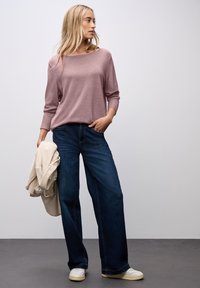 Maglione rosa chiaro a girocollo; vestibilità ampia. Jeans blu scuro a gamba larga; stile casual. Cardigan color crema tenuto in una mano. Sneakers bianche.