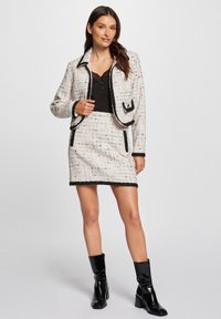 Blazer de tweed blanco con ribetes negros, combinado con una falda a juego. La modelo lleva una blusa negra y botas de tacón negro brillantes.