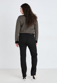 Pull en tricot avec un motif géométrique noir et doré, associé à un pantalon noir. Le modèle porte des escarpins beige à bout pointu, vu de dos.