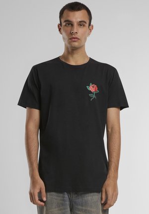 Jeune homme portant un t-shirt noir avec un petit motif de rose rouge sur la poitrine gauche, debout devant un fond uni gris clair.