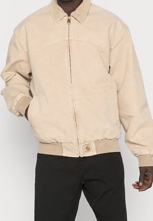 Personne portant une veste beige zippée avec poignets et ourlet côtelés, et un pantalon noir, debout avec une main dans la poche.