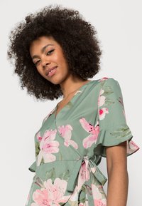 Vestido envelope floral em tecido verde, com flores rosas e brancas, mangas curtas com volantes e detalhe de cintura atada. Textura suave.