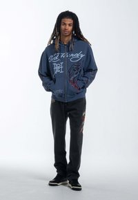 Hooded zip-up sweatshirt in navy with graphic prints, featuring a dragon design. Black denim jeans with colorful embroidery and black sneakers.