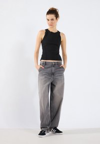 Svart tank topp med bølget hals, kombinert med løstsittende grå jeans. Modellen har på seg svart og hvite joggesko, står med hendene i lommene.