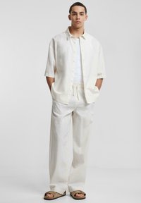 Witte linnen overhemd met korte mouwen, gecombineerd met losse witte linnen broek. Comfortabele bruine sandalen maken de outfit compleet.