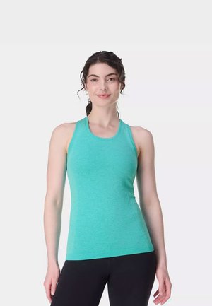 Jonge vrouw met krullend haar in een paardenstaart, gekleed in een turquoise mouwloze sporttop en zwarte legging, glimlachend naar de camera.