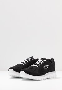Svarte joggesko med hvite såler og snorer, med en stilisert "S"-logo på siden, vist på en hvit overflate.