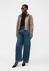 Γυναίκα με μπεζ τζάκετ με φερμουάρ, μαύρο ζιβάγκο, φαρδιά μπλε τζιν, μαύρη ζώνη, μαύρες μυτερές γόβες και γυαλιά ηλίου, στέκεται μπροστά σε λευκό φόντο.