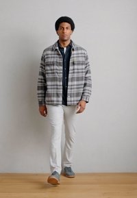 Γκρι πουκάμισο-τζάκετ με καρό σχέδιο και δύο τσέπες στο στήθος, φορεμένο πάνω από μπλε πουκάμισο, συνδυασμένο με λευκό παντελόνι και γκρι αθλητικά παπούτσια σε ξύλινο πάτωμα.