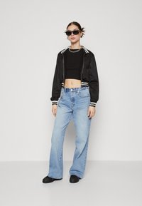 Chaqueta bomber corta negra con rayas blancas, top negro ajustado, jeans de pierna ancha en azul claro y zapatos negros sin cordones. Los complementos incluyen gafas de sol y un collar de cadena.