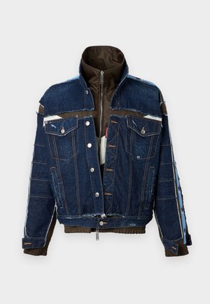 Joven con chaqueta de mezclilla oscura y acolchada en capas, camiseta blanca con diseño gráfico, pantalones negros y botas negras con cordones, de pie frente a un fondo blanco.