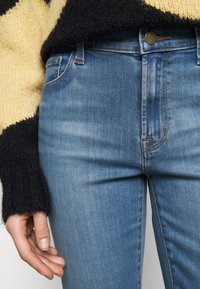 Jeans azules desgastados con corte slim y decoloraciones sutiles. Presentan un cierre frontal con botón y detalles en costura naranja. Suéter texturizado negro y amarillo.