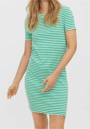Femme portant une robe à manches courtes rayée horizontale verte et blanche, debout devant un fond uni.