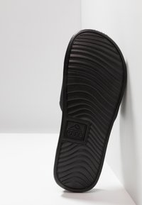 Semelle en caoutchouc noir avec des rainures texturées, arborant un logo en relief. Le bord du talon est arrondi et la forme générale est plate.