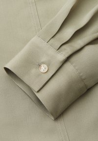 Nahaufnahme eines hellbeigen Hemdärmels mit einem einzelnen weißen Knopf und sichtbaren Nähten auf strukturiertem Stoff.